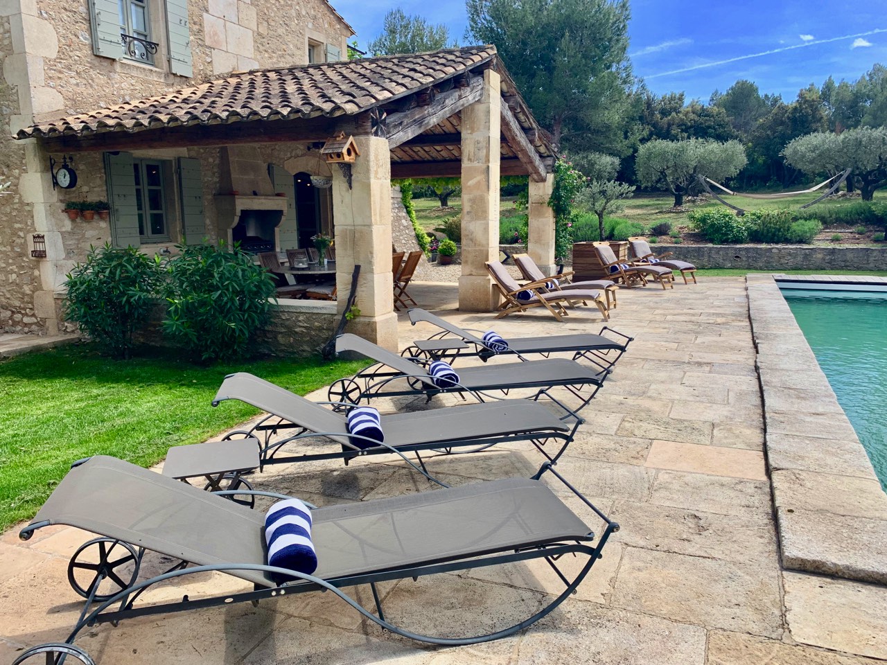 A serene poolside area at a rustic stone house, featuring lounging chairs under a pergola, surrounded by lush greenery and olive trees.